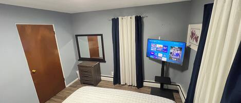 A bedroom with a bed, dresser, mirror, curtains, and a TV displaying a home screen.