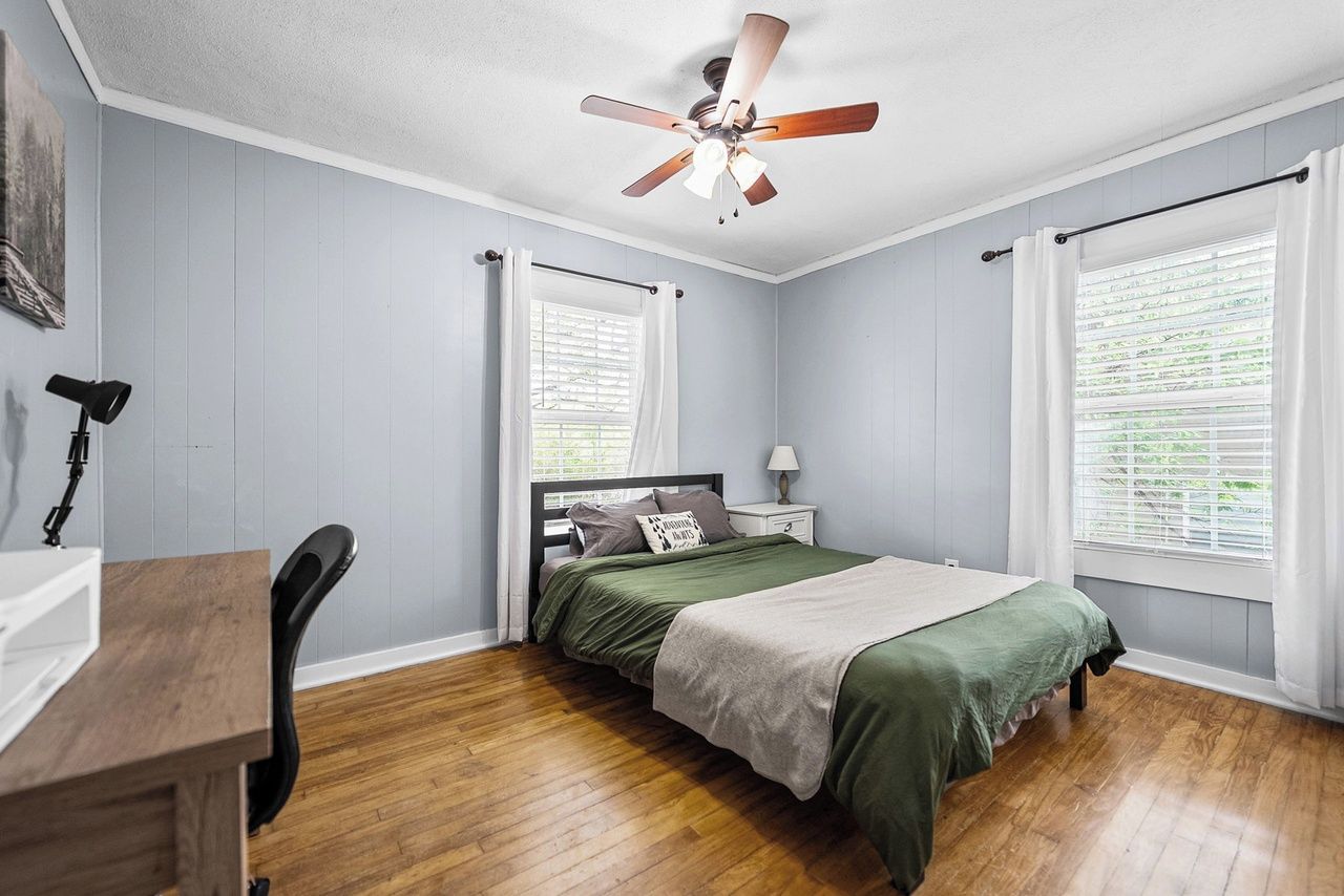 A bedroom with a bed, a desk, a chair, a ceiling fan, and windows with curtains.