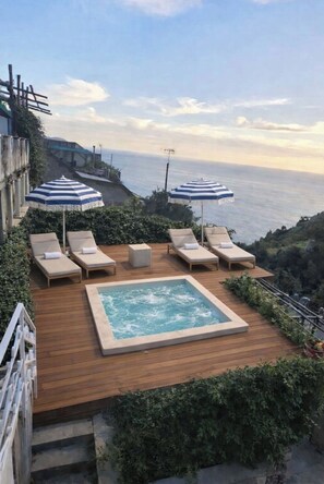 Eine Holzterrasse mit Whirlpool, Liegestühlen und Sonnenschirmen mit Blick aufs Meer.