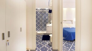 Ein schmaler Flur führt zu einem Badezimmer mit gemusterten Fliesen und einem Bett mit blauer Bettdecke.