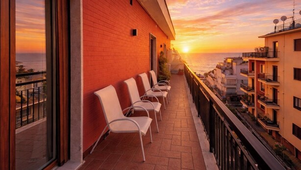 Ein Balkon mit weißen Stühlen, einer roten Backsteinmauer und Blick auf den Sonnenuntergang über dem Meer.