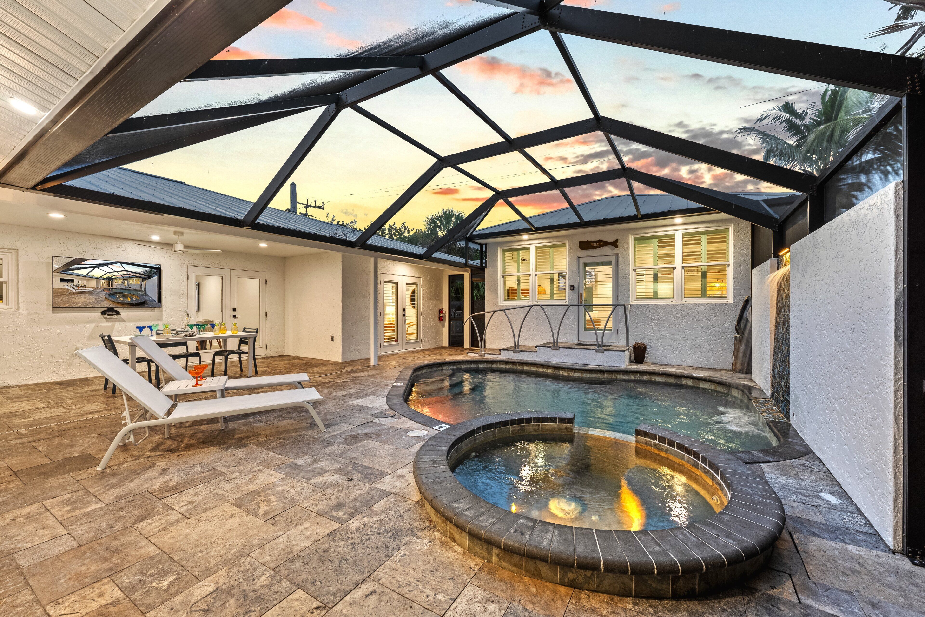 A covered outdoor swimming pool with a hot tub, a dining area, and a view of the sunset through a glass ceiling.