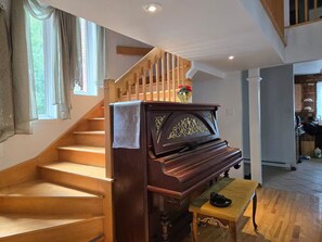 A grand piano in a spacious room with a wooden staircase, a small table, and a kitchen area in the background. - 6BedR with 7 beds Single house 3 private parking (Brossard)