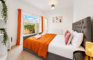 A bedroom with a large bed, a radiator, a window with curtains, a plant, and a wall-mounted artwork.