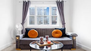 Una sala de estar moderna con un sofá gris, cojines naranja, una mesa de centro de vidrio y una ventana con cortinas.