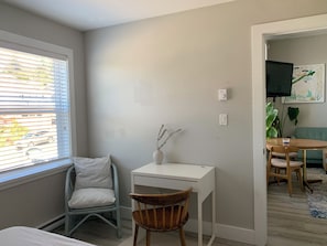 Une chambre à coucher comprenant un lit, un bureau, une chaise, une fenêtre avec des stores, un téléviseur fixé au mur et un canapé vert dans la pièce adjacente.