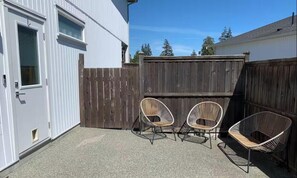 A patio with two wicker chairs and a table, a wooden fence, and a white building with a door and windows. - Sooke Broomhill Abode (Sooke)
