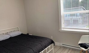 A bedroom with a bed, a chair, a window with blinds, and a radiator. - Sooke Broomhill Abode (Sooke)
