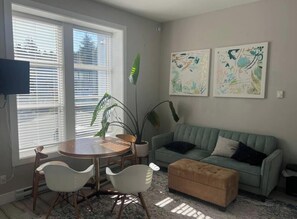 A living room with a round table, two white chairs, a green sofa, and a brown ottoman. There are two abstract paintings on the wall and a large plant. - Sooke Broomhill Abode (Sooke)