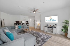 Ein modernes Wohnzimmer mit einer grauen Eckcouch, einem Deckenventilator, einem Essbereich mit Stühlen, einer Küchenzeile und einem großen Bildschirm, der 'HOST & HOME' anzeigt.