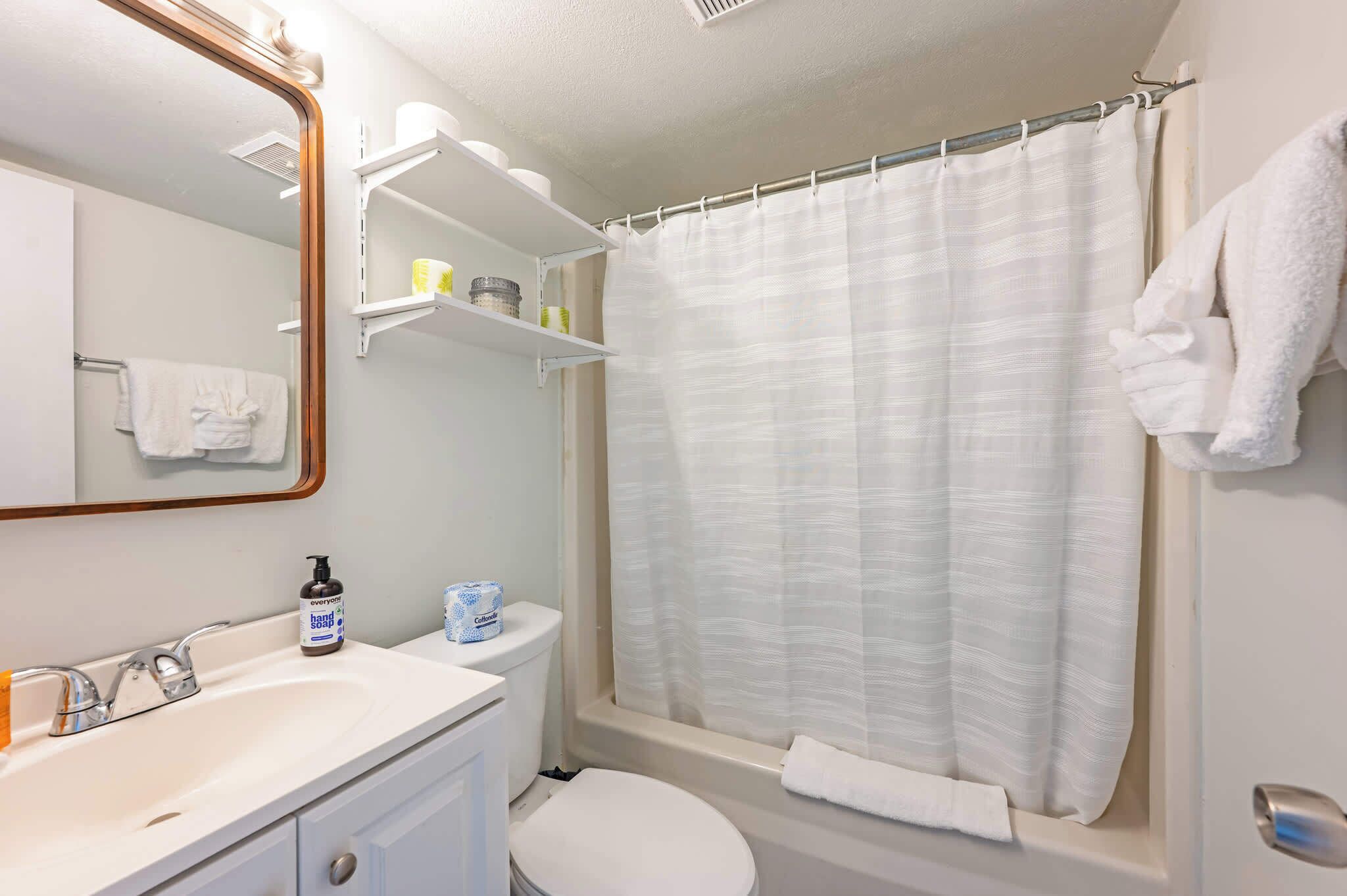 A bathroom with a white sink, toilet, and shower curtain. There are shelves above the sink with toiletries.