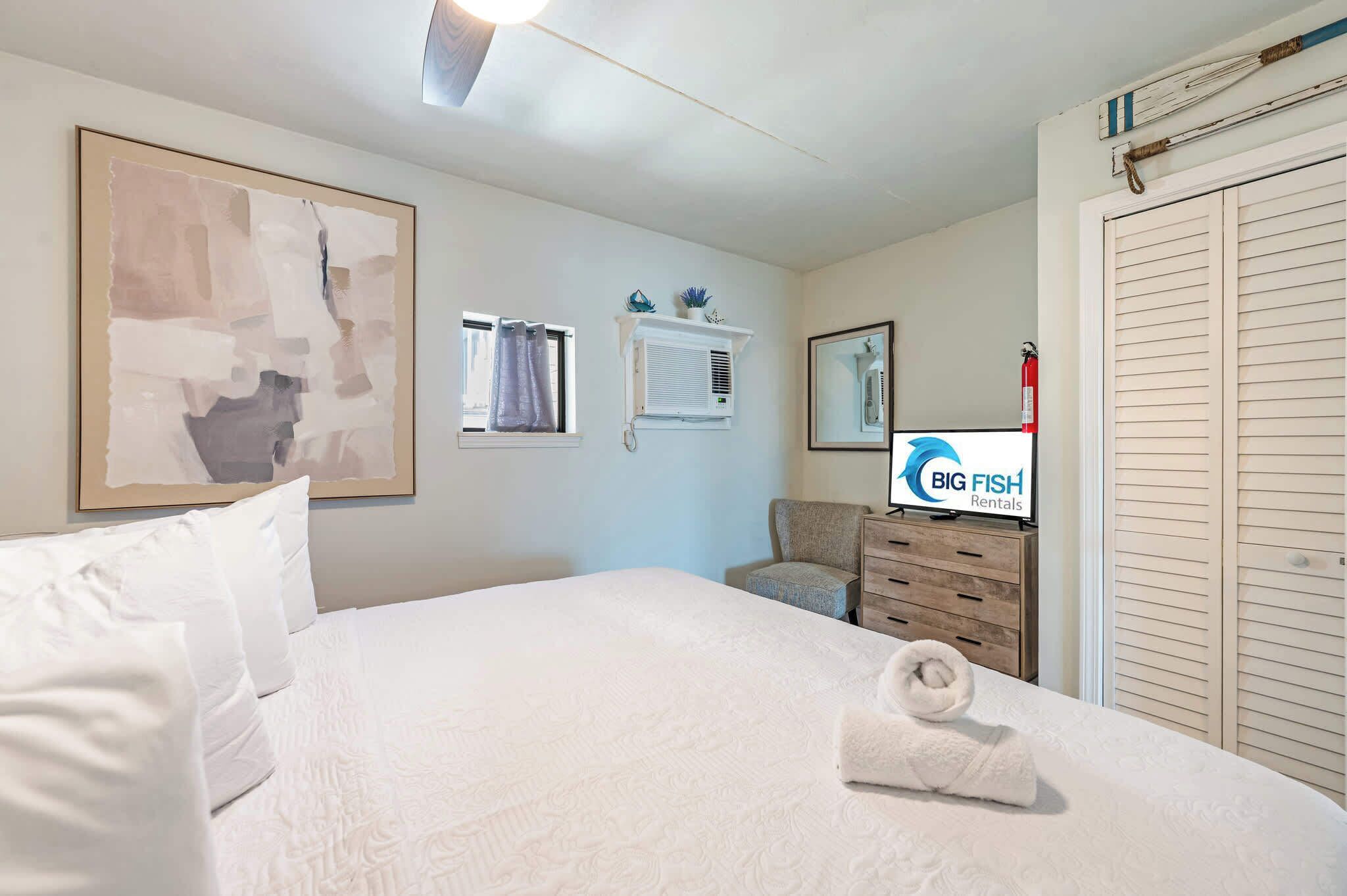 Une chambre d’hôtel comprenant un lit, une chaise, une commode, un climatiseur fixé au mur, un tableau encadré et un extincteur.