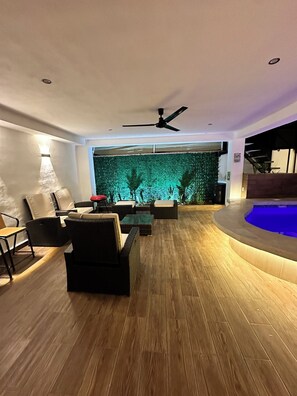 Ein modernes Wohnzimmer mit Holzboden, schwarzen Sofas, einem Deckenventilator und einer Wand mit einem grünen Blattmuster.