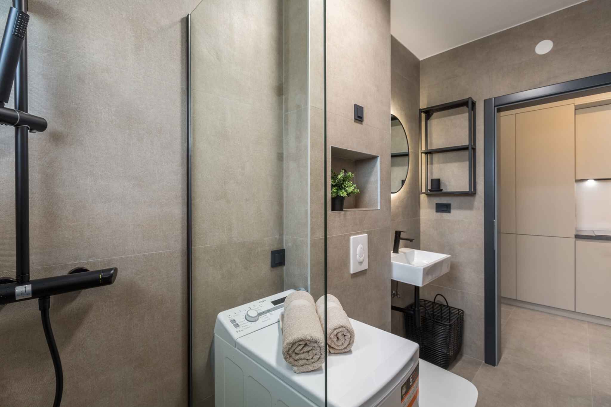 Une salle de bain moderne équipée d’un lave-linge intégré, d’un miroir, d’un lavabo et d’un espace douche.
