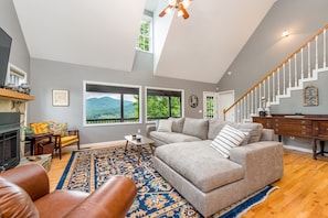 Un salon avec une cheminée, une grande fenêtre donnant sur les montagnes, un escalier et un coin repas.