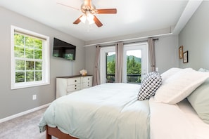 Une chambre à coucher comprenant un lit, un ventilateur de plafond, une commode, un téléviseur et une fenêtre donnant sur des arbres.