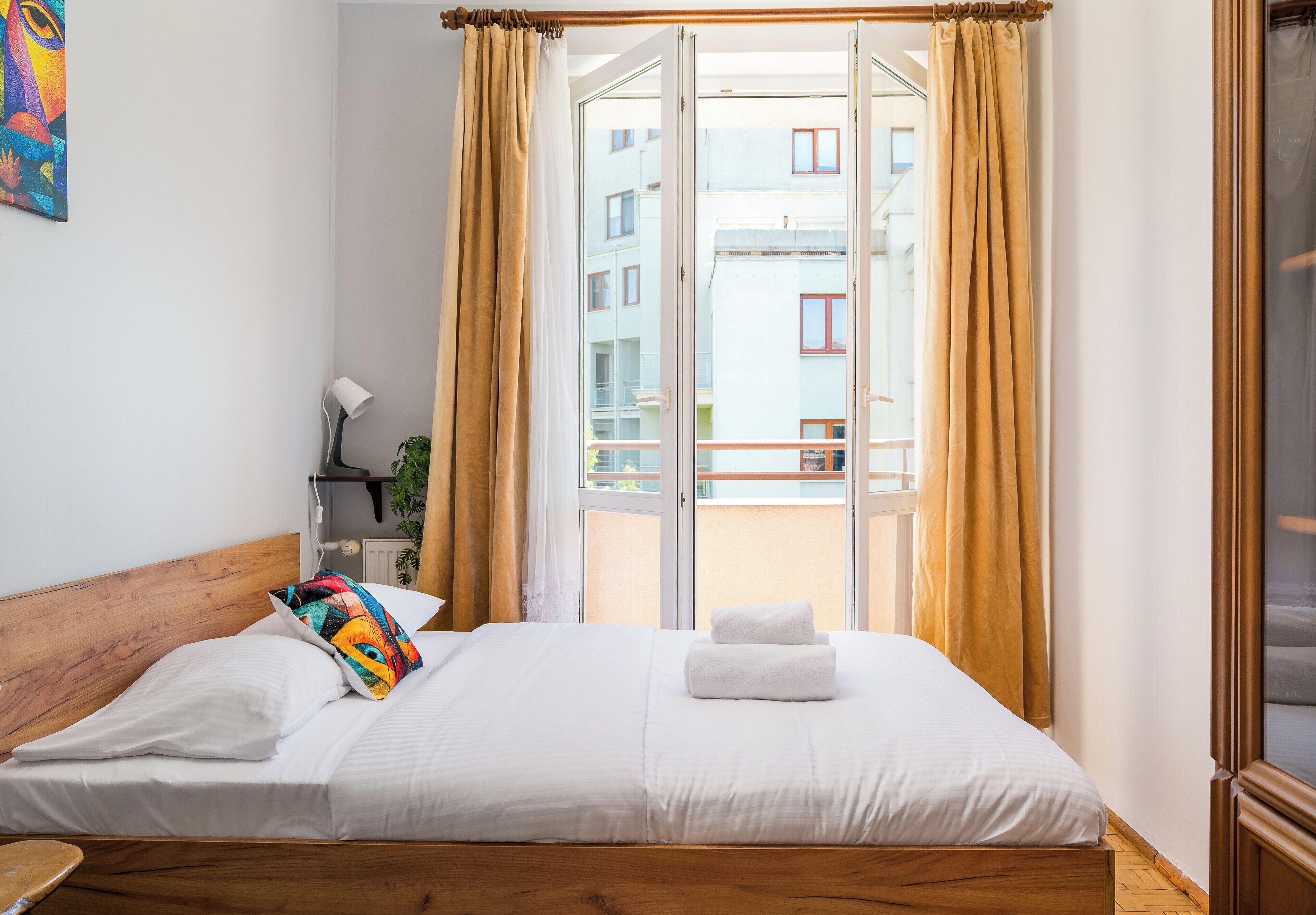 Une chambre d’hôtel avec une tête de lit en bois, un lit recouvert de draps blancs, un coussin coloré, une serviette blanche et une vue sur un immeuble à travers une porte vitrée.