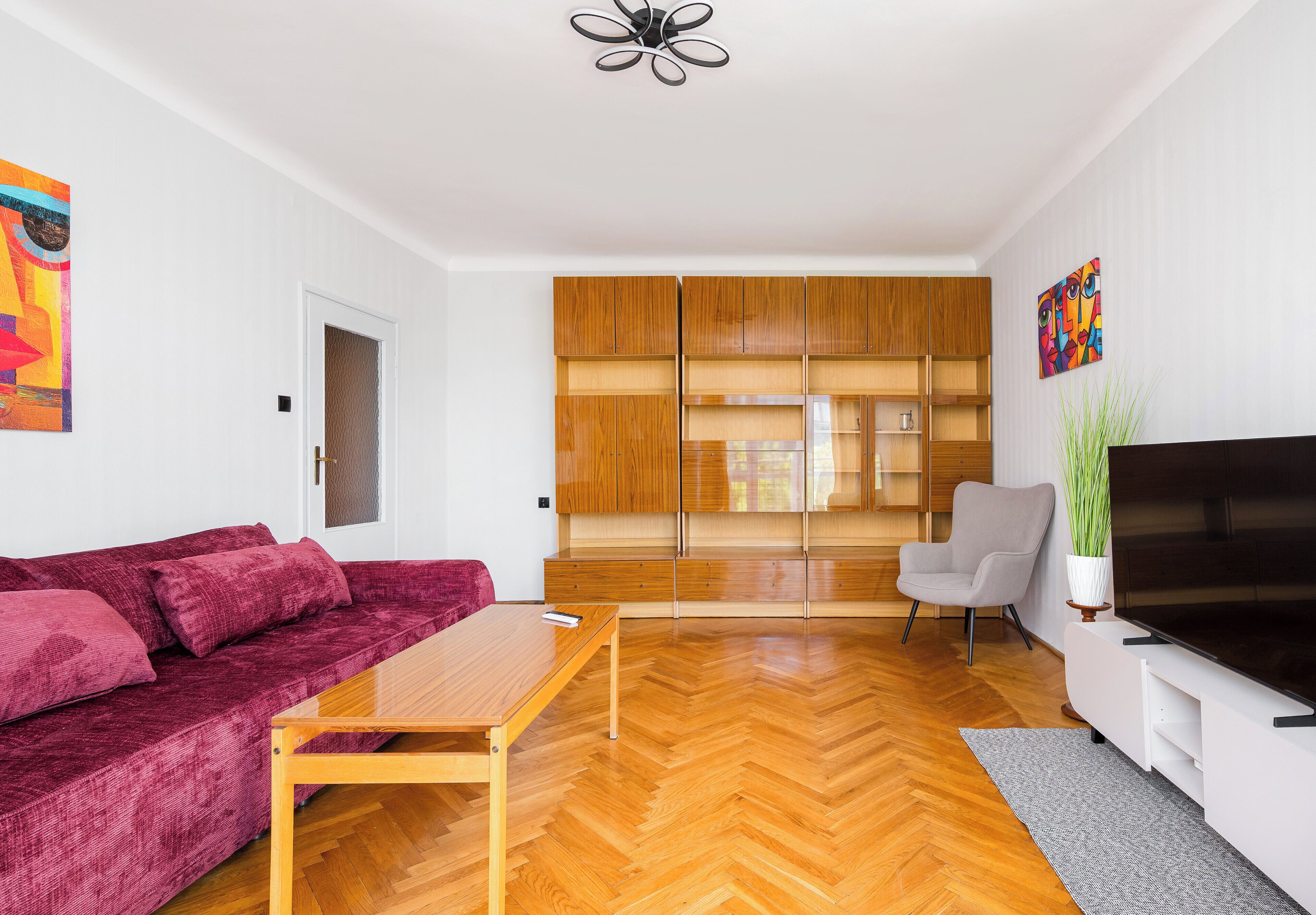 Un salon moderne avec un parquet en bois disposé en chevrons, un canapé rouge, une table basse en bois, un fauteuil gris, un téléviseur et un coin cuisine doté de meubles en bois.