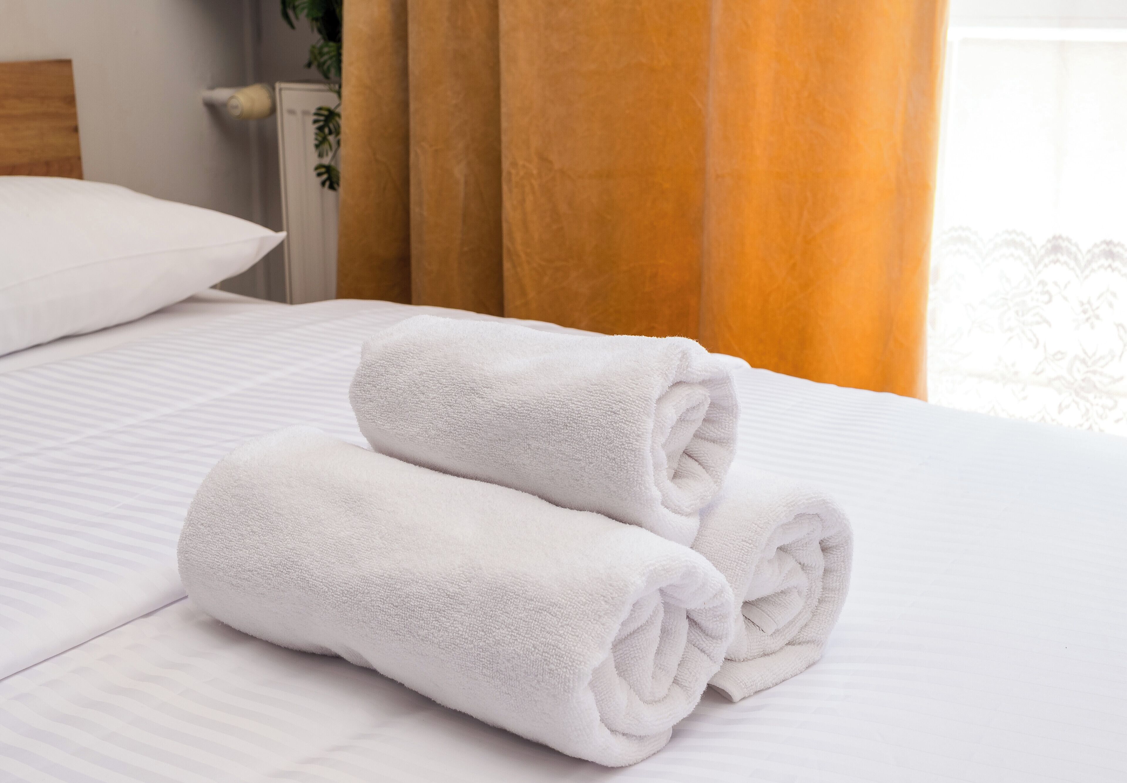 A neatly made bed with two rolled white towels, a white pillow, and a window with curtains.