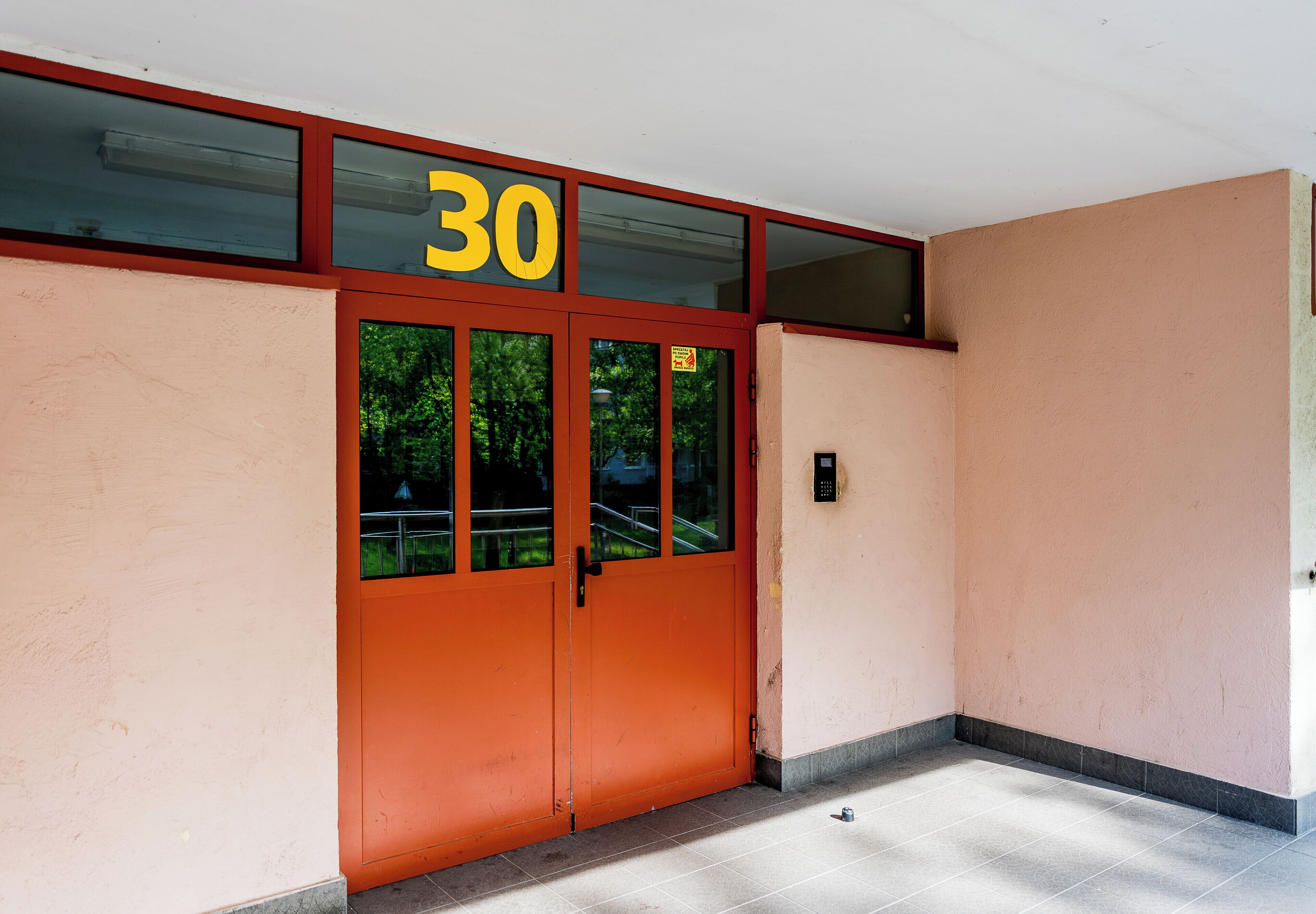 A building entrance with a red door, a number '30' above it, and a small electronic device on the wall.