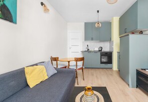 Espaço compacto com sofá, mesa de jantar, cadeiras, cozinha equipada com armários e eletrodomésticos, e um tapete pequeno.