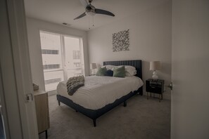 Un dormitorio con cama, ventilador de techo, mesita de noche con lámpara y ventana con persianas.