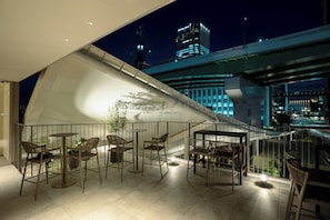 Een terras op het dak met zitplaatsen buiten en uitzicht op de skyline van de stad 's nachts.