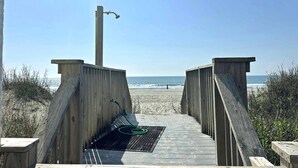 Een houten looppad dat naar een strand leidt, met een metalen rooster en een groene slang op de grond.