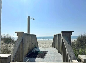 Ein Holzsteg führt zu einem Strand, wo ein Metallgitter und ein grüner Schlauch am Boden liegen.