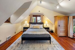 Un dormitorio con techo inclinado, una cama con cabecera, dos mesitas de noche con lámparas, un armario de madera y una ventana con cortinas.