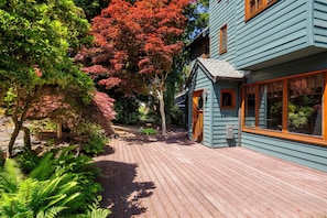Una terraza de madera que conduce a una casa con porche, rodeada de abundante vegetación y árboles con hojas rojas.