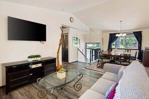 A modern living room with a flat-screen TV, a glass coffee table, a sofa, a dining area with a table and chairs, and a staircase leading to another level.
