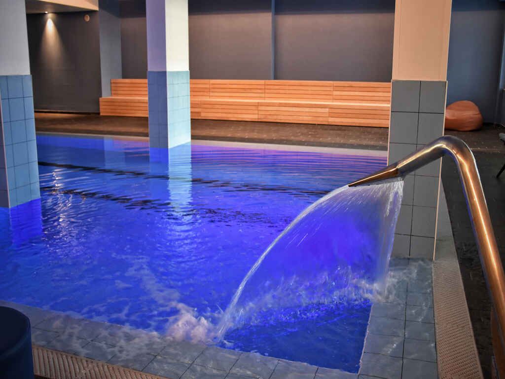 Piscina interior com um elemento aquático, paredes azulejadas e assentos de madeira.