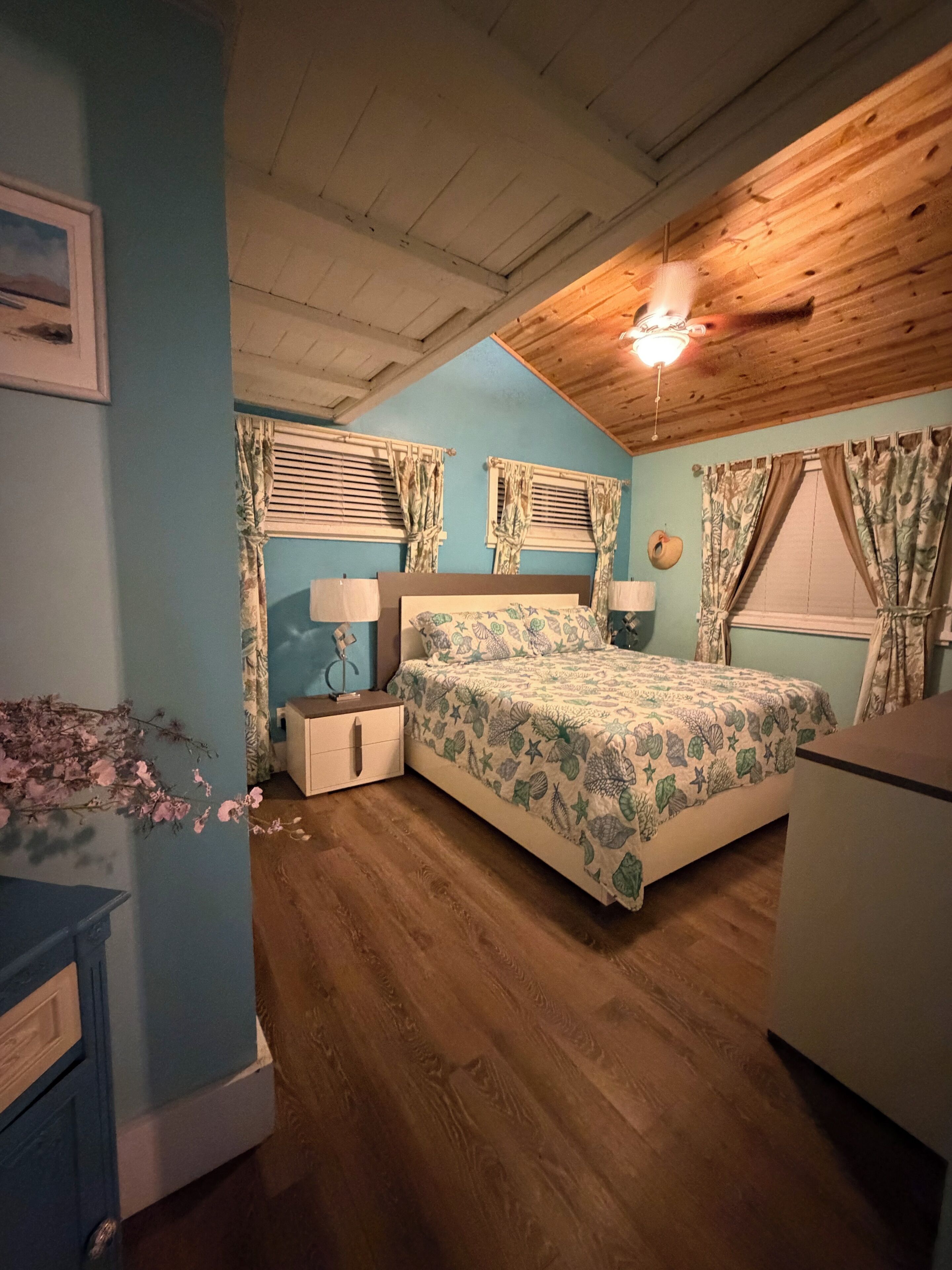 A bedroom with a wooden ceiling, a bed with floral bedding, two bedside tables with lamps, and a dresser.