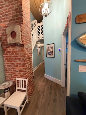 A narrow hallway with a brick wall, a wooden staircase, a white chair, a small table, and a framed picture on the wall. - Casa Verano, a cozy 2-bedroom bungalow with loft in charming Panama City Beach! (Panama City Beach)