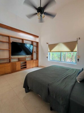 A bedroom with a bed, a TV on a wooden shelf, a ceiling fan, and a window with a view of greenery. - Beautiful house 5 min from the hotel zone (Cancún)