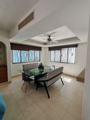 A dining area with a glass table, two chairs, a ceiling fan, and barred windows. - Beautiful house 5 min from the hotel zone (Cancún)