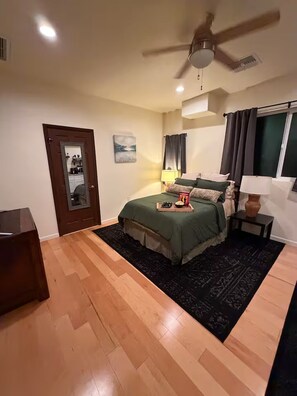 Un dormitorio con una cama, ventilador de techo, ventana con cortinas, mesita de noche y espejo.