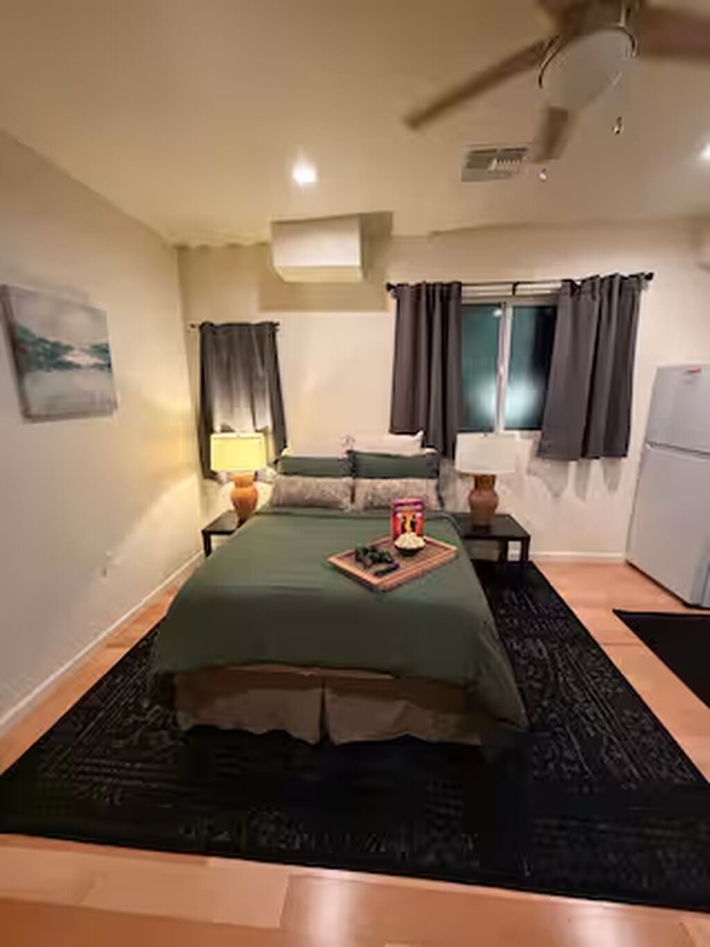 A room with a bed, a nightstand, a lamp, a window with curtains, a wall-mounted air conditioner, and a small table with a book.