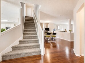 Eine Treppe führt zu einer zweiten Etage, ein Küchenbereich mit weißen Schränken und ein Essbereich mit zwei Stühlen.
