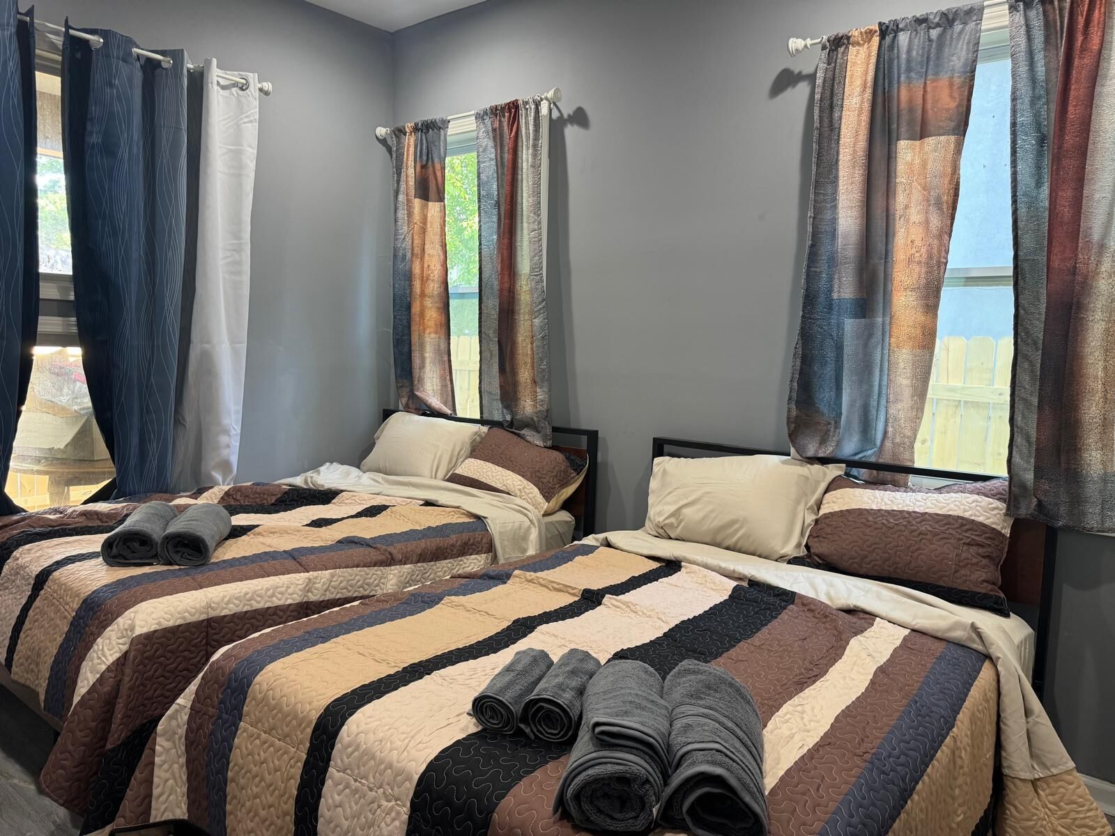 A bedroom with two beds, a window with curtains, and rolled towels on the bed.