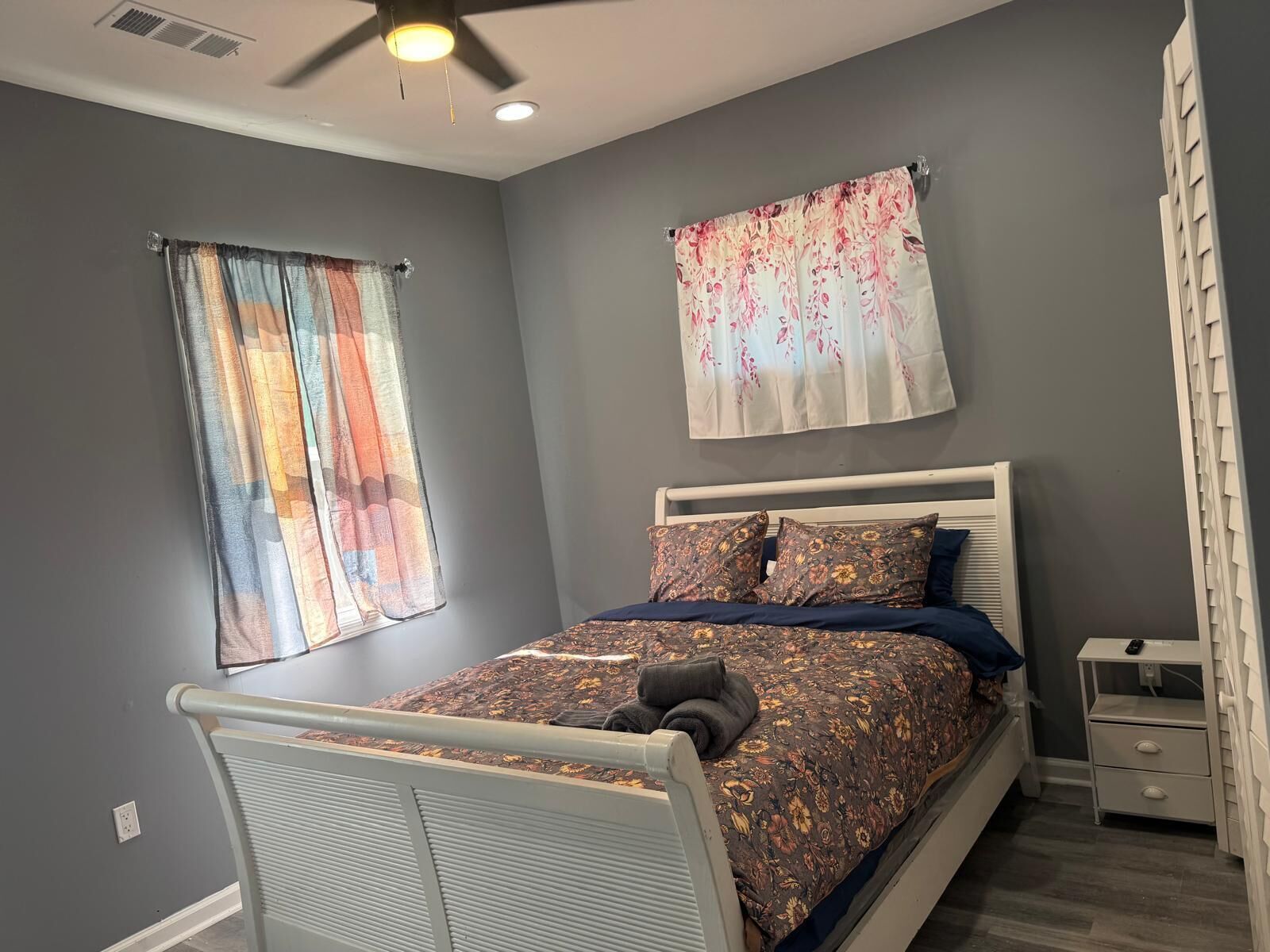A bedroom with a bed, two curtains, a ceiling fan, and a nightstand.