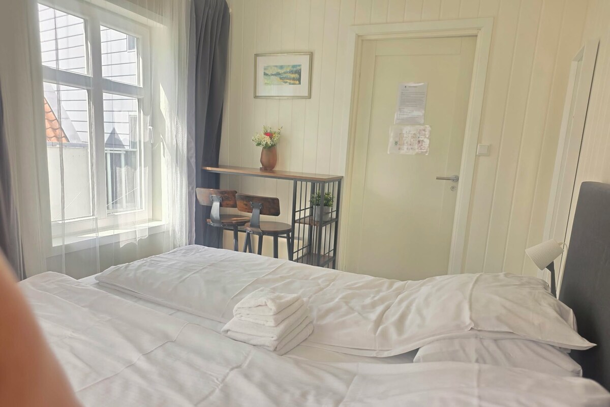 A hotel room with a bed, a desk with a chair, a small table with a vase, and a door with a notice.