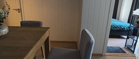 A room with a wooden dining table, a grey cushioned chair, a framed picture on the wall, and a view of a balcony with a bed.