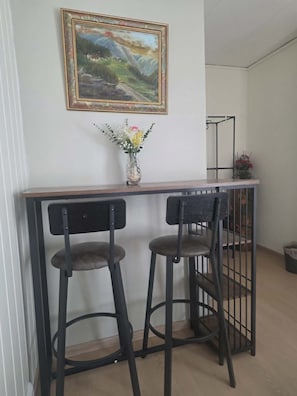 A dining area with a wooden table, two black barstools, a vase with flowers, and a framed painting on the wall.