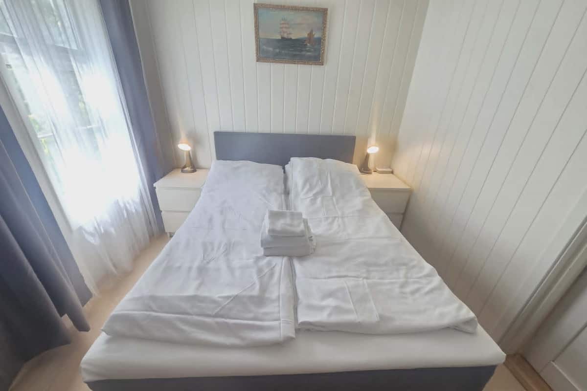 A neatly made bed with white linens, two bedside lamps, a framed picture on the wall, and a window with curtains.