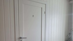A white door with the number 2, a small table with a vase, and a chair in a room with white paneled walls. - Stavangerliving (Stavanger)