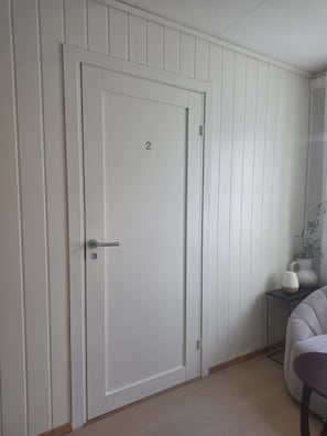 A white door with the number 2, a small table with a vase, and a chair in a room with white paneled walls.