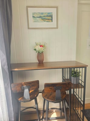 A small dining area with a wooden table, two chairs, a vase with flowers, and a framed painting on the wall.