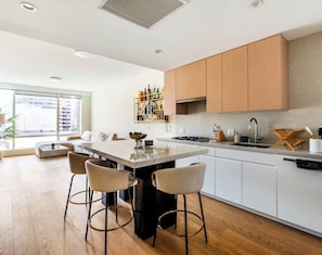 A modern kitchen with a central island, bar stools, wooden cabinets, and a large window. - Elevated City Living - Resort Amenities, 24/7 Security, Free Parking, Gym, Sauna (Los Angeles)
